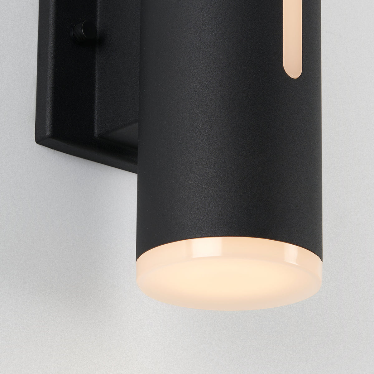 Close-up of a modern black cylindrical wall light fixture mounted on a light-colored wall, emitting a soft, warm glow from the frosted bottom and a slim vertical cutout.