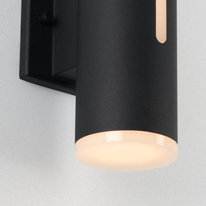 A close-up of the Artika Linea modern indoor-outdoor LED wall light, featuring a black cylindrical design with a frosted bottom diffuser, emits a soft, warm glow while mounted on a light-colored wall.