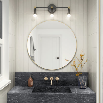 Modern bathroom with a round mirror above a black marble sink, brass fixtures, brown soap dispenser, glass vase with yellow branches, and the Artika Wayden LED mid-century bathroom vanity light (3 CCT). White walls and door reflected in mirror.