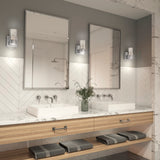 Modern bathroom with two square sinks, marble countertop, wood shelves with rolled towels, two large mirrors, wall sconces, and a vase with greenery. White herringbone tile backsplash and a walk-in shower are visible.