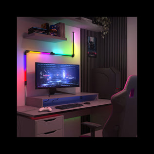 A modern gaming setup features an Artika Fitch smart foldable RGB LED gaming floor lamp beside a desk with a monitor displaying a futuristic game, white controller, keyboard, and mouse. A gaming chair and wall poster complete the scene.