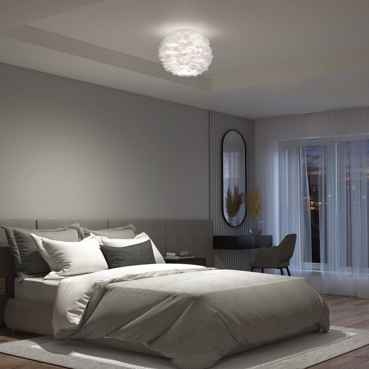 Modern bedroom with a gray bed, layered bedding, a decorative ceiling light fixture, a desk with a chair, a tall oval mirror, and a window with sheer curtains letting in evening light.