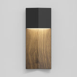 A modern wall sconce with a black top section and a wooden lower section, emitting a warm light downward onto the wood grain surface, mounted on a light gray wall.