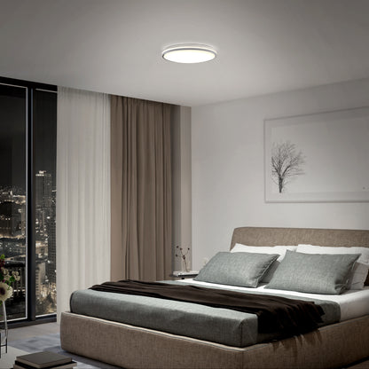 Modern bedroom with a large bed, gray bedding, and pillows. The Artika Macy 15 in. LED modern flush mount light (RGB Wifi) is on. Floor-to-ceiling window shows a night cityscape; neutral curtains and tree artwork decorate the wall.