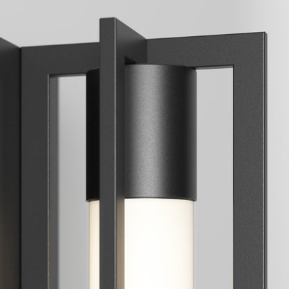 Close-up of the Artika Walt indoor-outdoor LED wall light, featuring a black metal frame and cylindrical frosted glass shade, emitting soft white light against a light gray background.