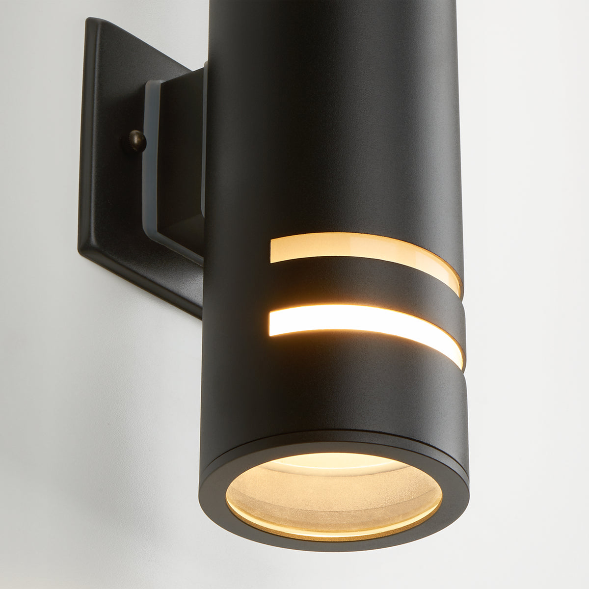 A modern black cylindrical wall sconce with two horizontal light slots emits warm light, mounted on a square black base against a white wall.