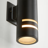 A modern black cylindrical wall sconce with two horizontal light slots emits warm light, mounted on a square black base against a white wall.