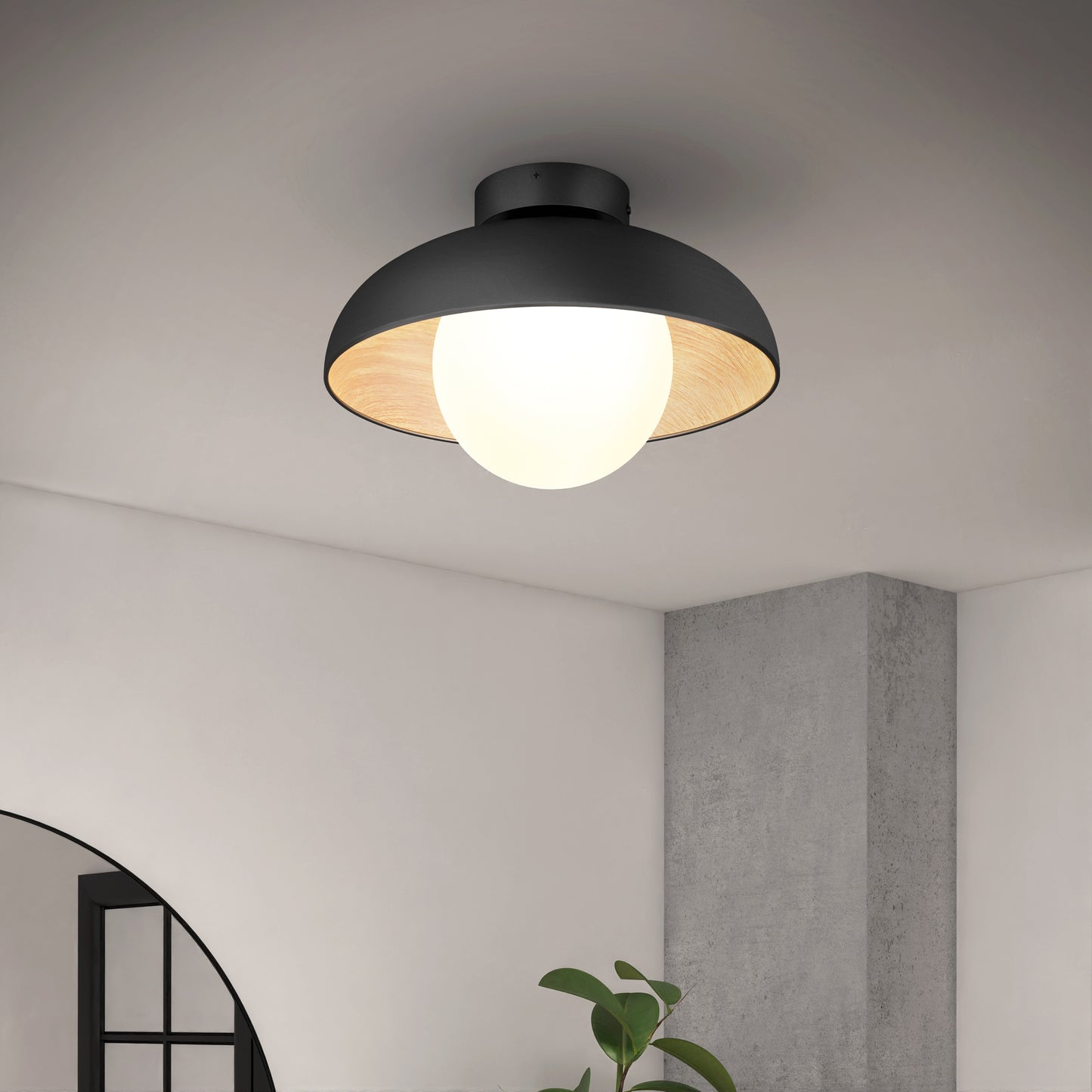 The Artika Boden LED flush mount light 5 CCT is a modern black ceiling fixture with a large exposed bulb, shown mounted on a white ceiling in a contemporary room with a plant, window, and concrete column visible below.
