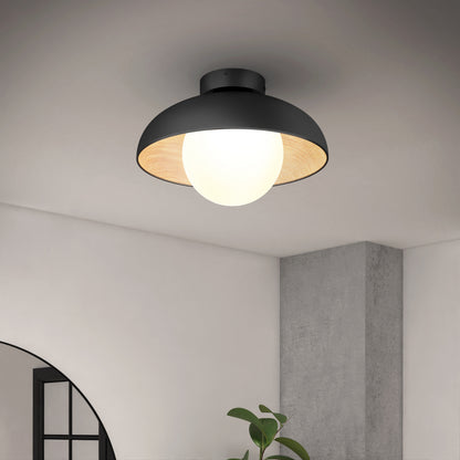 The Artika Boden LED flush mount light 5 CCT is a modern black ceiling fixture with a large exposed bulb, shown mounted on a white ceiling in a contemporary room with a plant, window, and concrete column visible below.