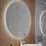 A round, backlit mirror is mounted on a wooden slat wall in a modern bathroom. Below the mirror is a white sink with a silver faucet, a soap dispenser, and a small vase with green foliage.