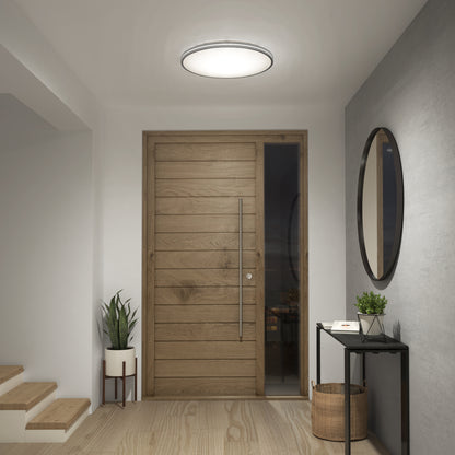 A modern entryway features a wooden front door, black console table with decor, round mirror, white potted plant, wooden stairs on the left, and an Artika Athos 21 in. LED flush mount light with remote overhead.