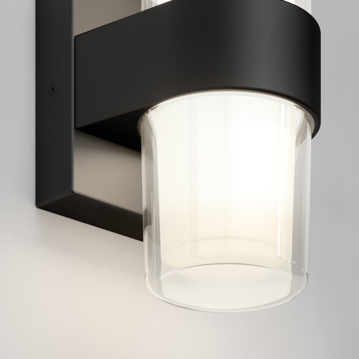 A close-up of a modern wall-mounted light fixture with a black metal frame and a cylindrical frosted glass shade, illuminating a soft white light against a light-colored wall.