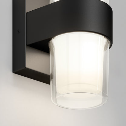A close-up of the Colton modern LED indoor-outdoor wall light 5 CCT by Artika, featuring a cylindrical frosted glass shade and sleek black metal frame, illuminated against a light-colored wall.
