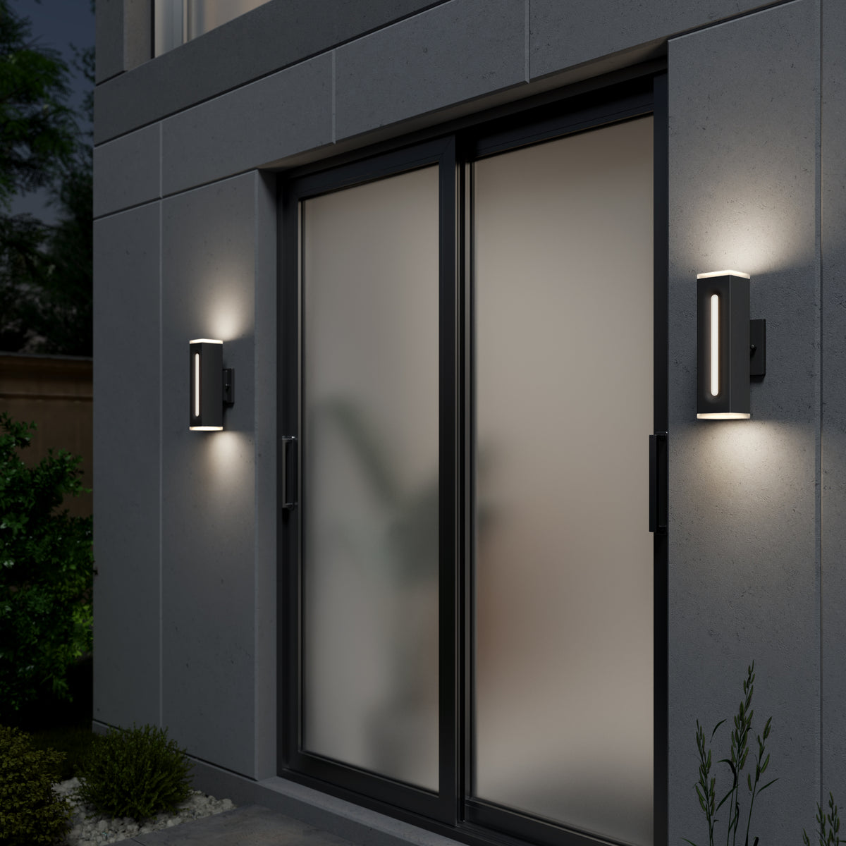 Modern wall lights illuminate both sides of a glass sliding door with frosted panels on a gray concrete building exterior, surrounded by plants and greenery at dusk.