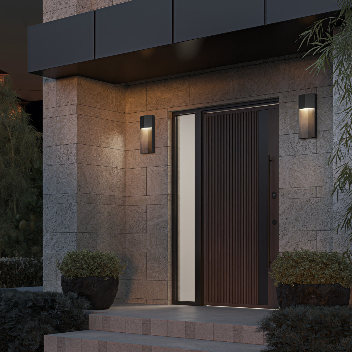 A modern stone house entrance with a dark wooden door, vertical frosted glass panel, two wall-mounted lights, stone steps, and potted plants on each side of the doorway.
