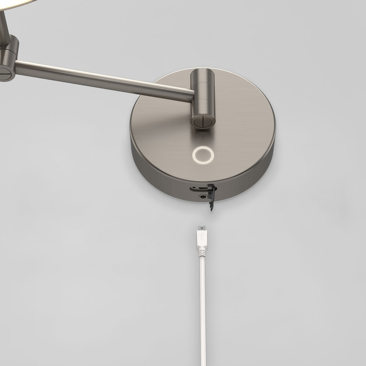A close-up of a modern wall-mounted lamp with a metallic finish, featuring a circular base and a USB cable connected to a port underneath the base against a plain light gray wall.