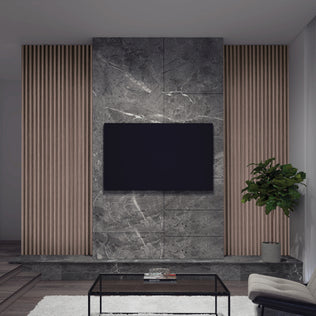 A modern living room wall features a mounted flat-screen TV, gray marble paneling, and Artika’s Panolok 30.67 ft wall panel kit (pack of 8) in oak finish as vertical slat panels, plus a potted plant, glass coffee table, and white Scandinavian rug.