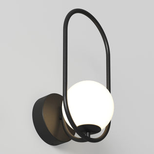 The Artika Bloom LED Sconce 3 CCT features a spherical white glass shade and matte black metal frame forming an elegant oval loop, mounted on a circular base against a plain light gray wall.
