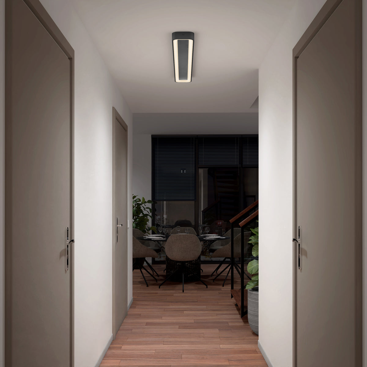 A hallway with wooden flooring and closed doors on both sides leads to a dining area with a round table, chairs, indoor plants, and large windows with dark blinds. A modern ceiling light illuminates the space.