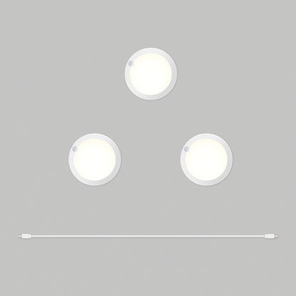 Three round white Artika Accent under cabinet LED light 3CCT puck lights are arranged in a triangle on a gray background, with a white USB cable positioned horizontally below them.