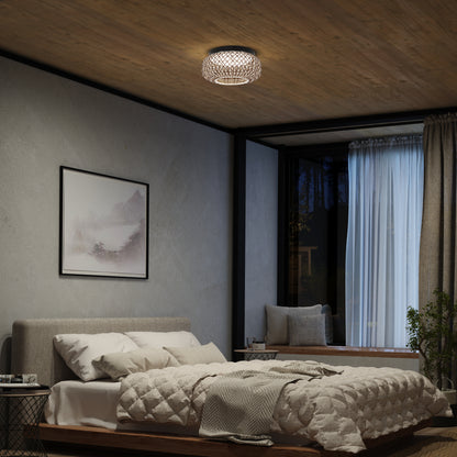 A cozy bedroom with a gray upholstered bed, textured bedding, a landscape painting, a large window with sheer curtains, and an Artika Benson 15 in. LED flush mount light with 5 CCT on the ceiling.