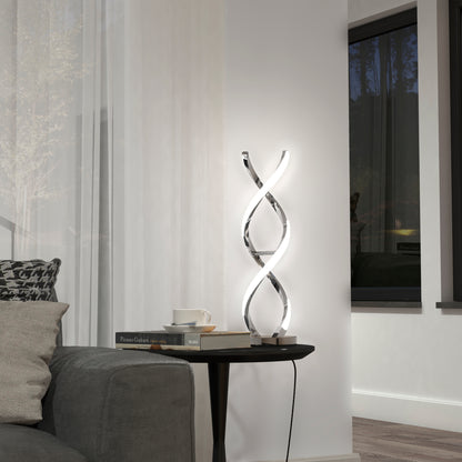 The Artika Swirl LED table lamp with a chrome finish sits on a small black side table beside a gray sofa, accompanied by books and a cup. A large window with sheer white curtains is in the background.