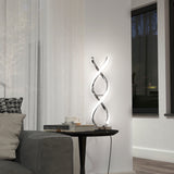 A modern living room features a gray sofa, a black side table with books and a cup, and a sculptural LED lamp with twisting light tubes. A large window and sheer white curtains are in the background.