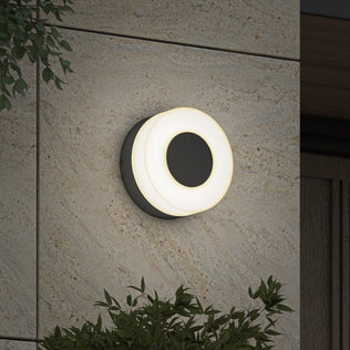 The Artika Ludwig modern LED indoor-outdoor wall light 5CCT, featuring a glowing white ring, is mounted on a textured stone exterior wall and surrounded by green leafy plants.