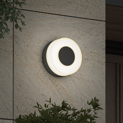 The Artika Ludwig modern LED indoor-outdoor wall light 5CCT, featuring a glowing white ring, is mounted on a textured stone exterior wall and surrounded by green leafy plants.