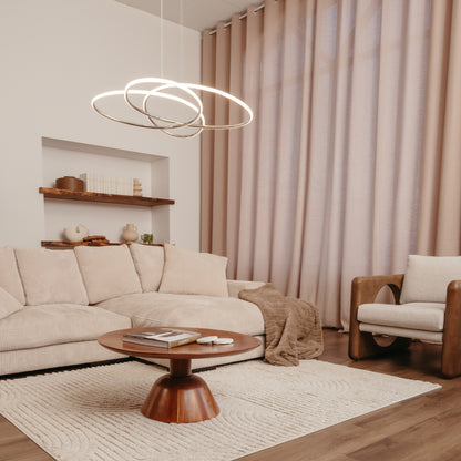A modern living room with beige furniture, a round wooden coffee table, plush rug, and soft floor-to-ceiling curtains. Overhead hangs the Artika Swirl Butterfly LED contemporary pendant light 3 CCT with circular rings.