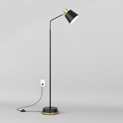 The Artika Alton LED modern floor lamp, featuring a sleek black design with gold accents, stands on a gray floor. Plugged into a white outlet on a light gray wall, the lamp is turned on and emits a gentle light.