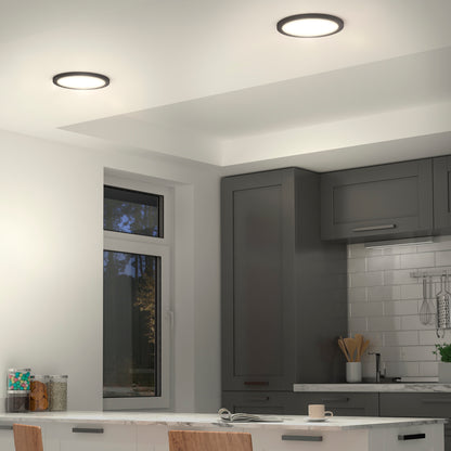 A modern kitchen features Artikas Europa Disk 9 in. LED flush mount lights, gray cabinets, a window, white subway tile backsplash, and a white countertop with a mug and plate. Wooden chairs are partially visible in the foreground.