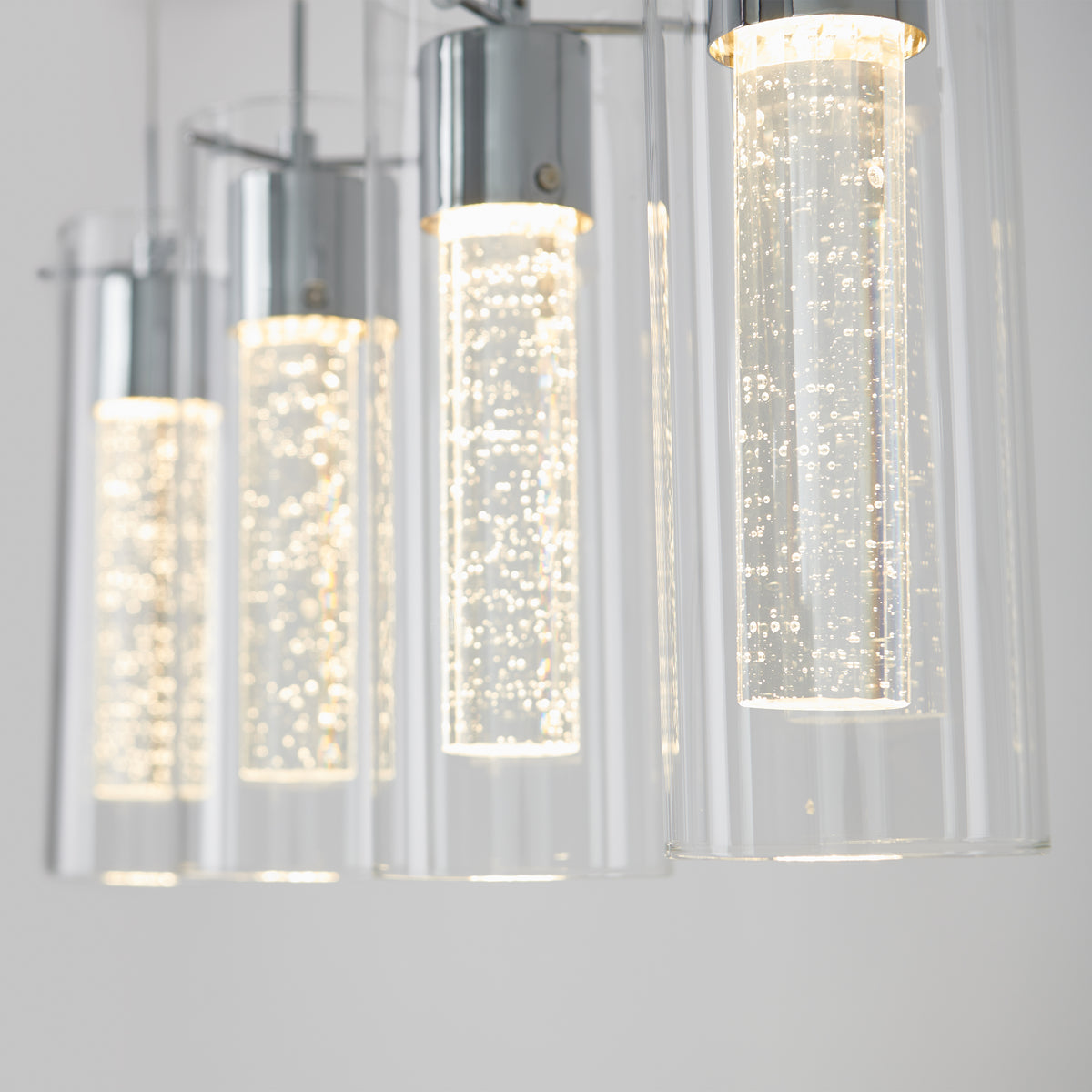 Close-up of modern hanging pendant lights with clear cylindrical glass shades, illuminated and featuring bubbles inside the glass, against a light, neutral background.