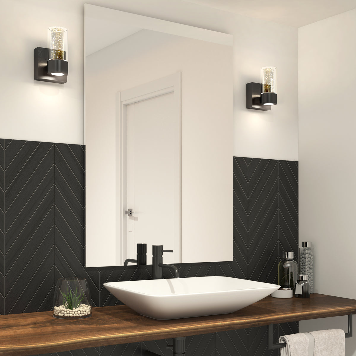 Modern bathroom with a large mirror, black chevron wall tiles, vessel sink on a wooden countertop, black faucet, wall-mounted lights, and various toiletries and decor items. Door reflected in the mirror.