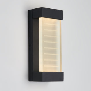 The Artika Percy LED indoor-outdoor wall light features a modern black rectangular frame and a frosted glass panel with horizontal etched lines, softly glowing against a plain light gray wall.