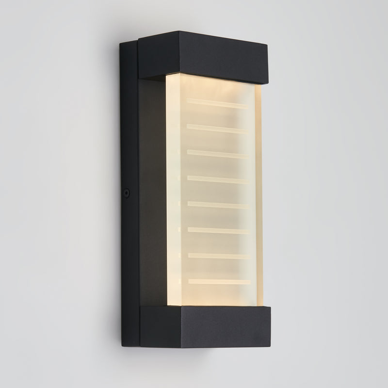 The Artika Percy LED indoor-outdoor wall light features a modern black rectangular frame and a frosted glass panel with horizontal etched lines, softly glowing against a plain light gray wall.