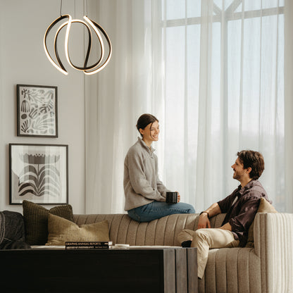 A woman and a man sit on a beige sofa in a cozy, modern living room with large windows, abstract wall art, and an Artika Vivaldi Big LED pendant light. The woman holds a mug and smiles at the man.