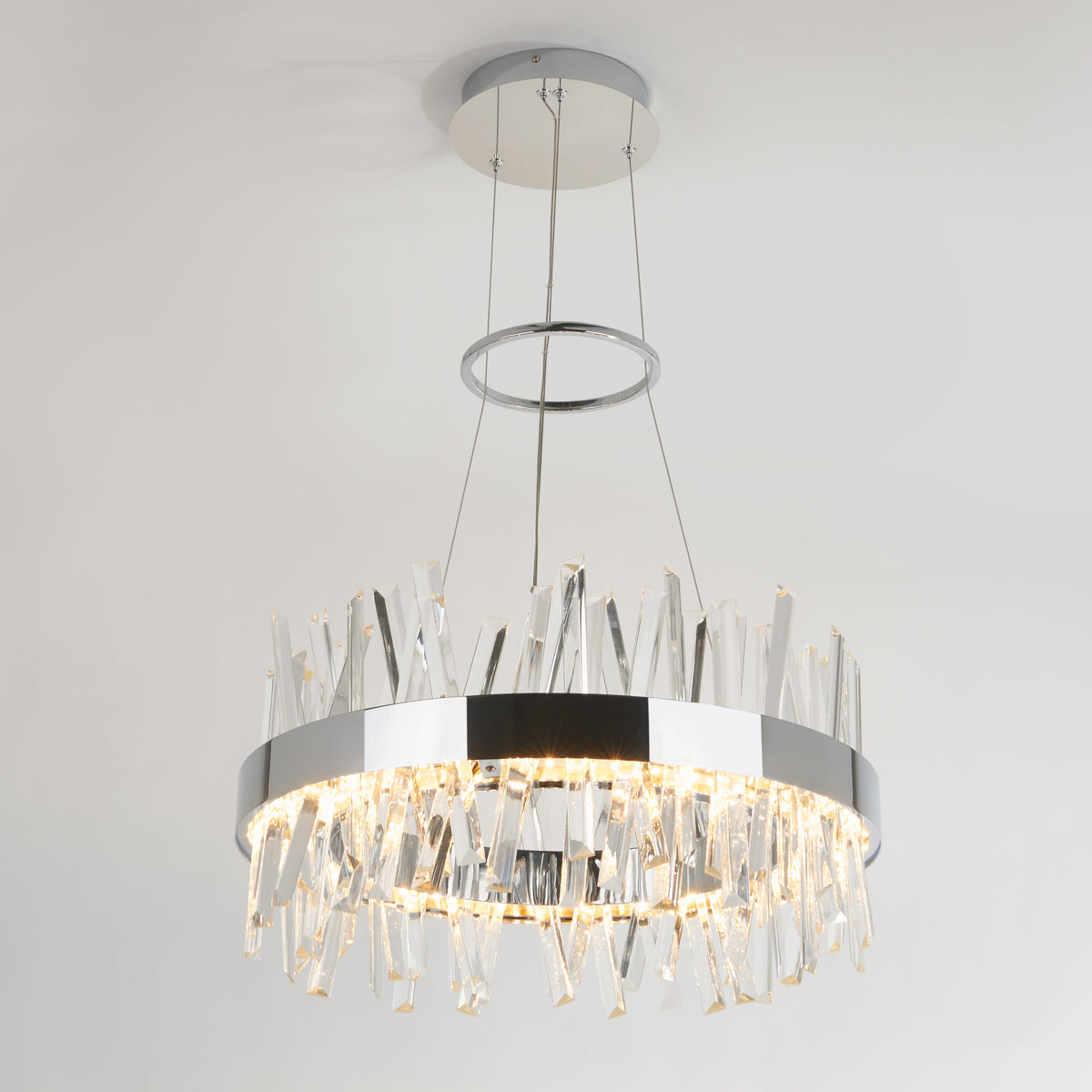 The Artika Enchanted LED pendant light is a modern circular chandelier with a chrome finish, suspended by cables and adorned with glass crystal prisms around the rim, casting radiant reflections on the white ceiling.