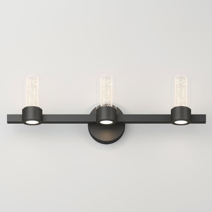 The Artika Novello modern LED bathroom vanity light features three sparkling textured cylindrical glass shades on a horizontal black bar with a circular base, all mounted on a white wall.
