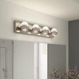 A modern bathroom light fixture with four spherical glass bulbs on a rectangular metal base is mounted above a mirror, with a window and decorative plants visible in the background.