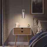 A modern bedroom nightstand with angled wooden legs holds a small plant, a geometric ornament, and a decorative object. A pendant light hangs above, casting a warm glow. A bed and framed art are partially visible nearby.
