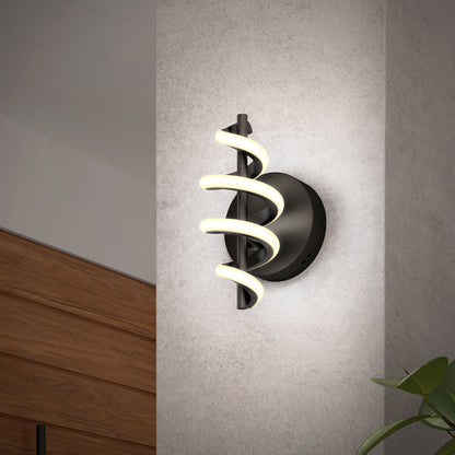 The Artika Chorus LED Wall Light features a modern black base with three spiral-shaped LED tubes, mounted on a light gray wall. A wooden cabinet and green plant are visible nearby.