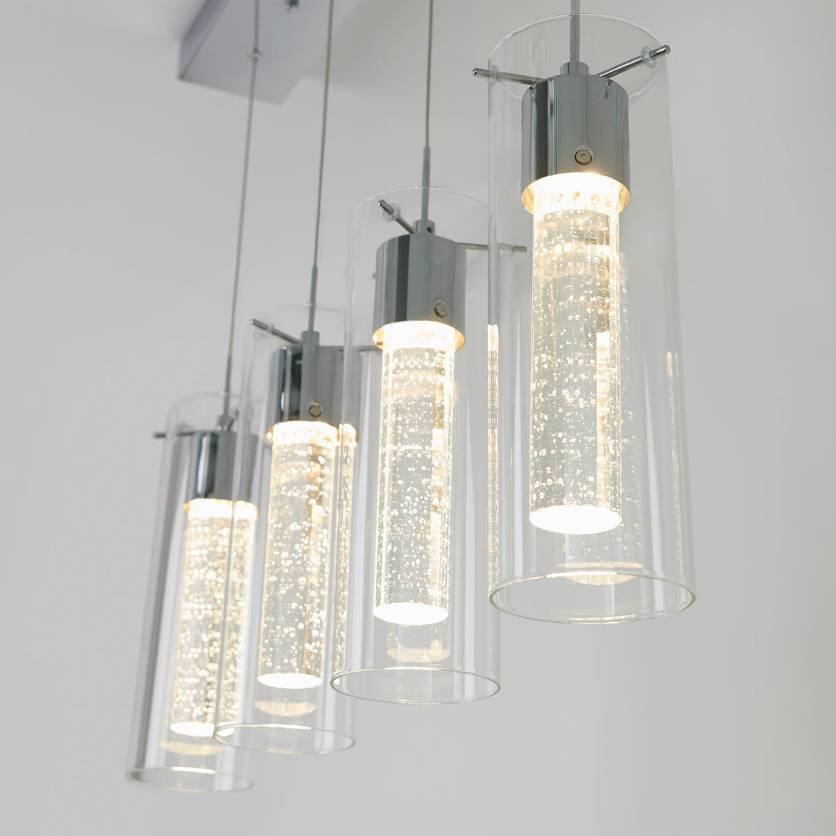 The Artika Essence 4-light LED pendant features four modern cylindrical lights with clear glass and chrome accents. Each light’s inner tube has small bubbles, creating a sparkling effect against a white background.