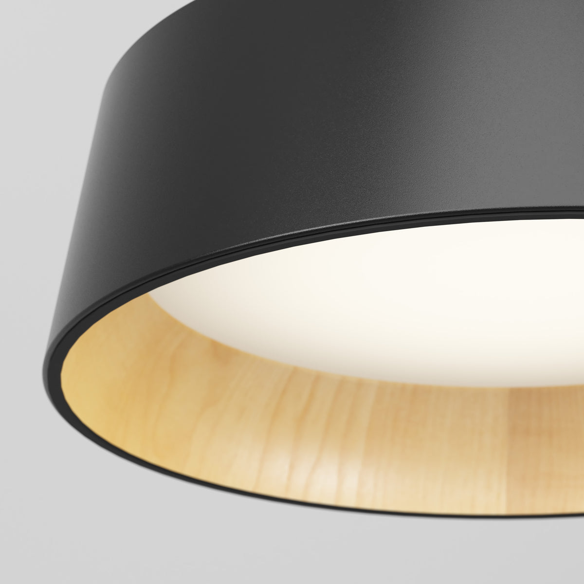 Close-up of a modern ceiling lamp with a black outer shade, light wood interior, and a glowing white light diffuser at the center, set against a plain light gray background.