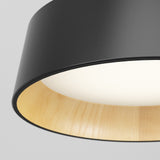Close-up of a modern ceiling lamp with a black outer shade, light wood interior, and a glowing white light diffuser at the center, set against a plain light gray background.