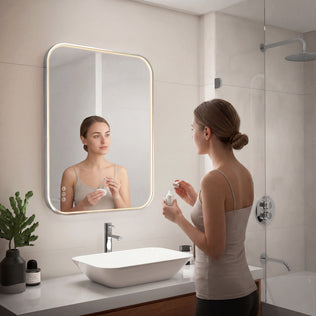 A woman stands at a modern sink, facing the Artika Royce Dimmable LED Mirror (3 CCT, horizontal/vertical mount), applying skincare. The bathroom has a glass shower, white tiles, and a potted plant on the counter.