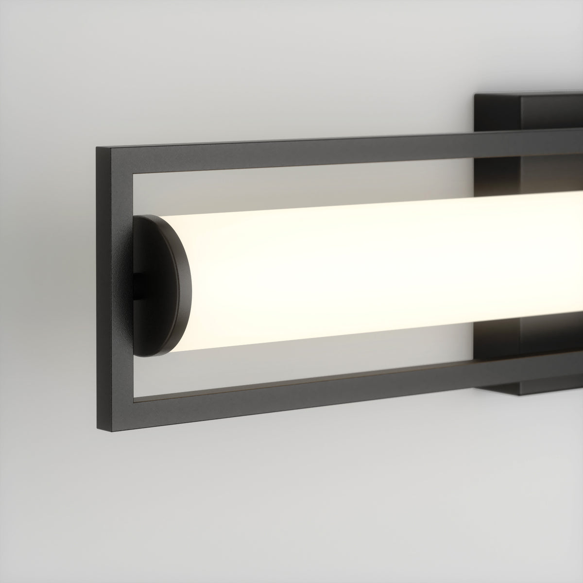 A modern wall-mounted light fixture with a rectangular black metal frame surrounding a cylindrical frosted glass light, emitting a soft white glow against a plain white wall.