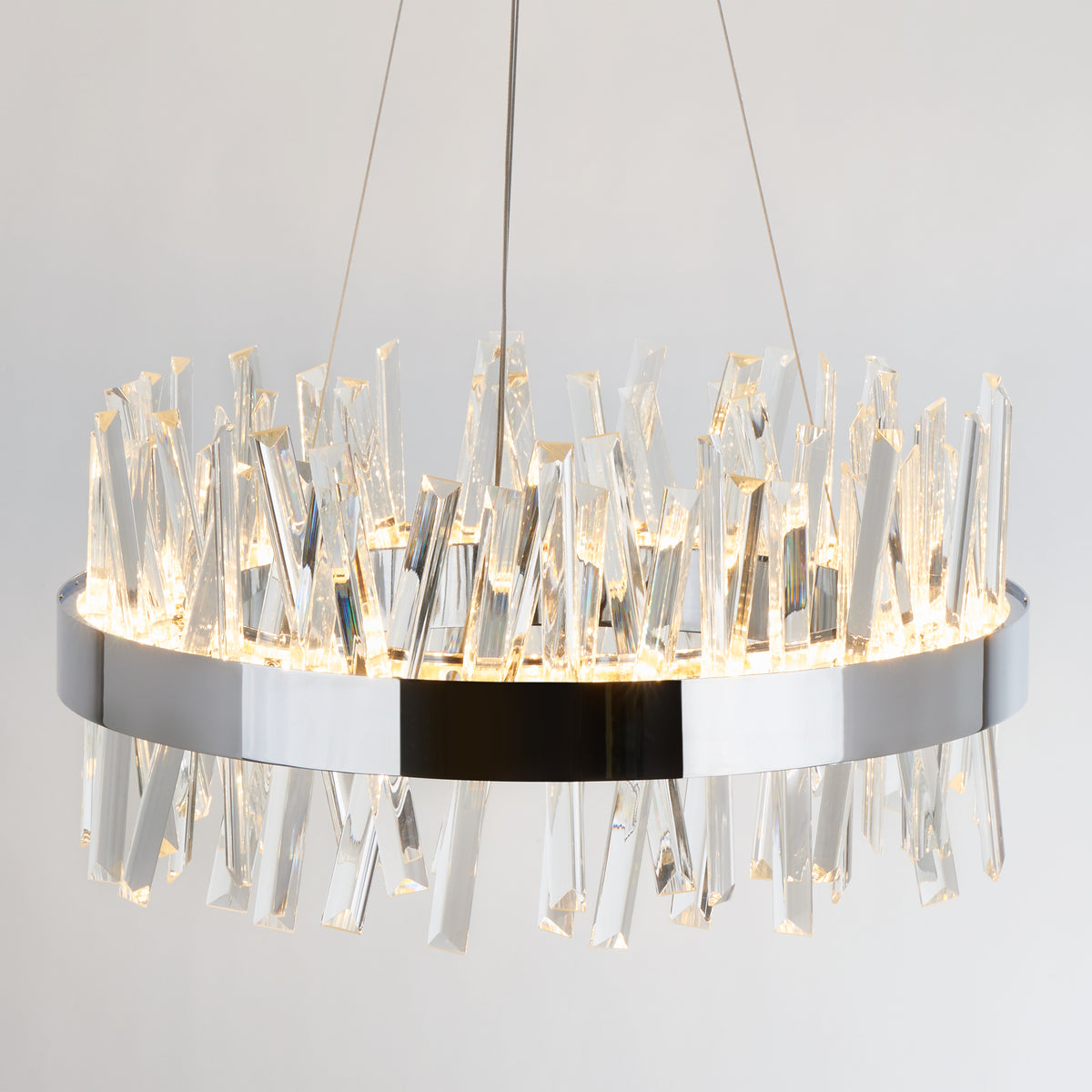 A modern circular chandelier with a metallic frame, featuring numerous vertical crystal prisms that reflect and scatter light, suspended by thin wires against a light background.