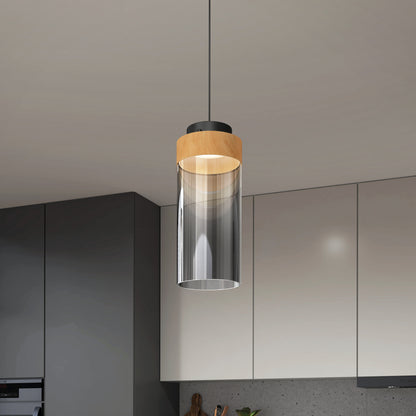 The Artika Hanson 1-light LED pendant with a cylindrical glass shade and wooden accent hangs from the ceiling in a contemporary kitchen with light and dark cabinets.