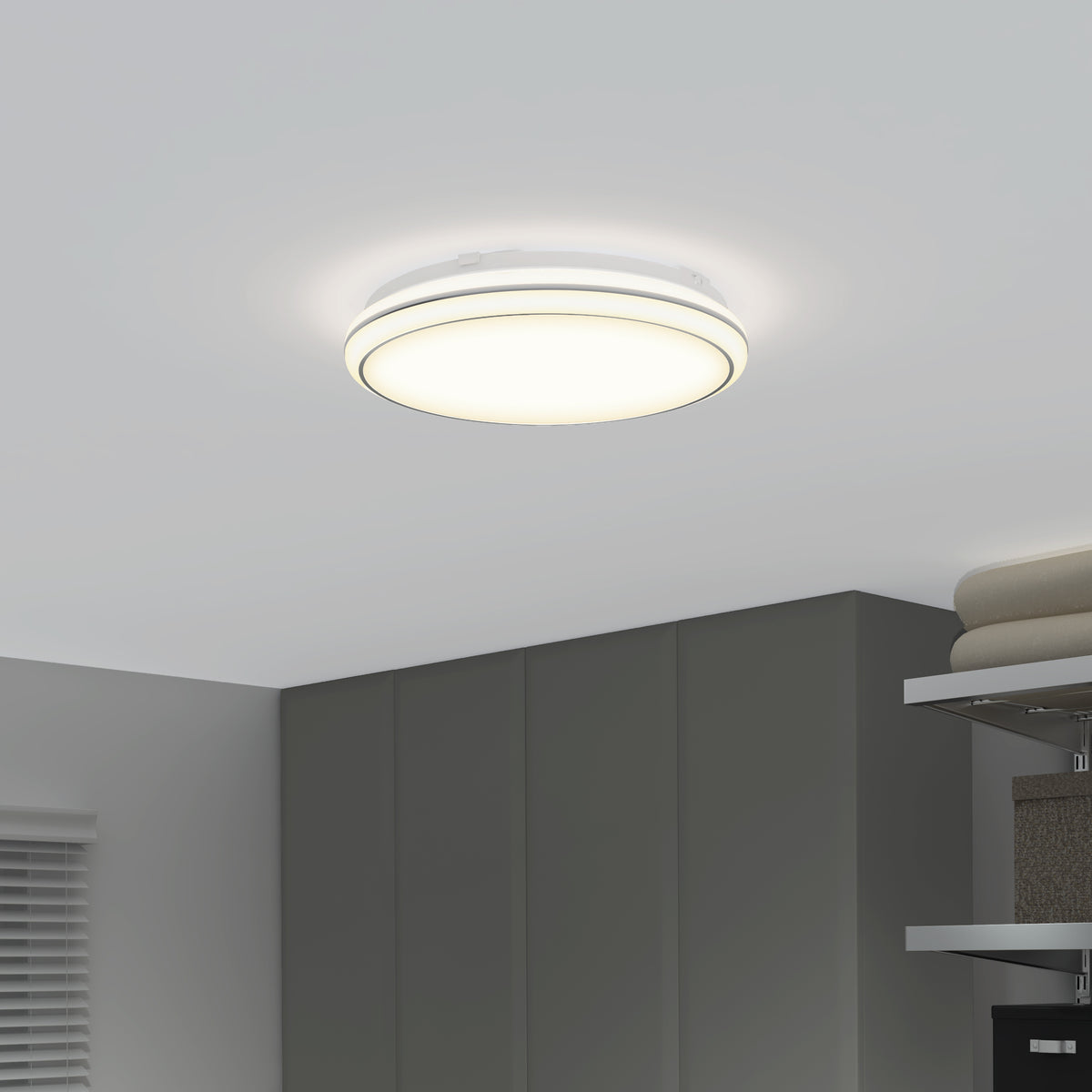 A modern, round ceiling light fixture is illuminated on a white ceiling above a minimalist room with gray cabinets, folded towels, and a window with blinds.